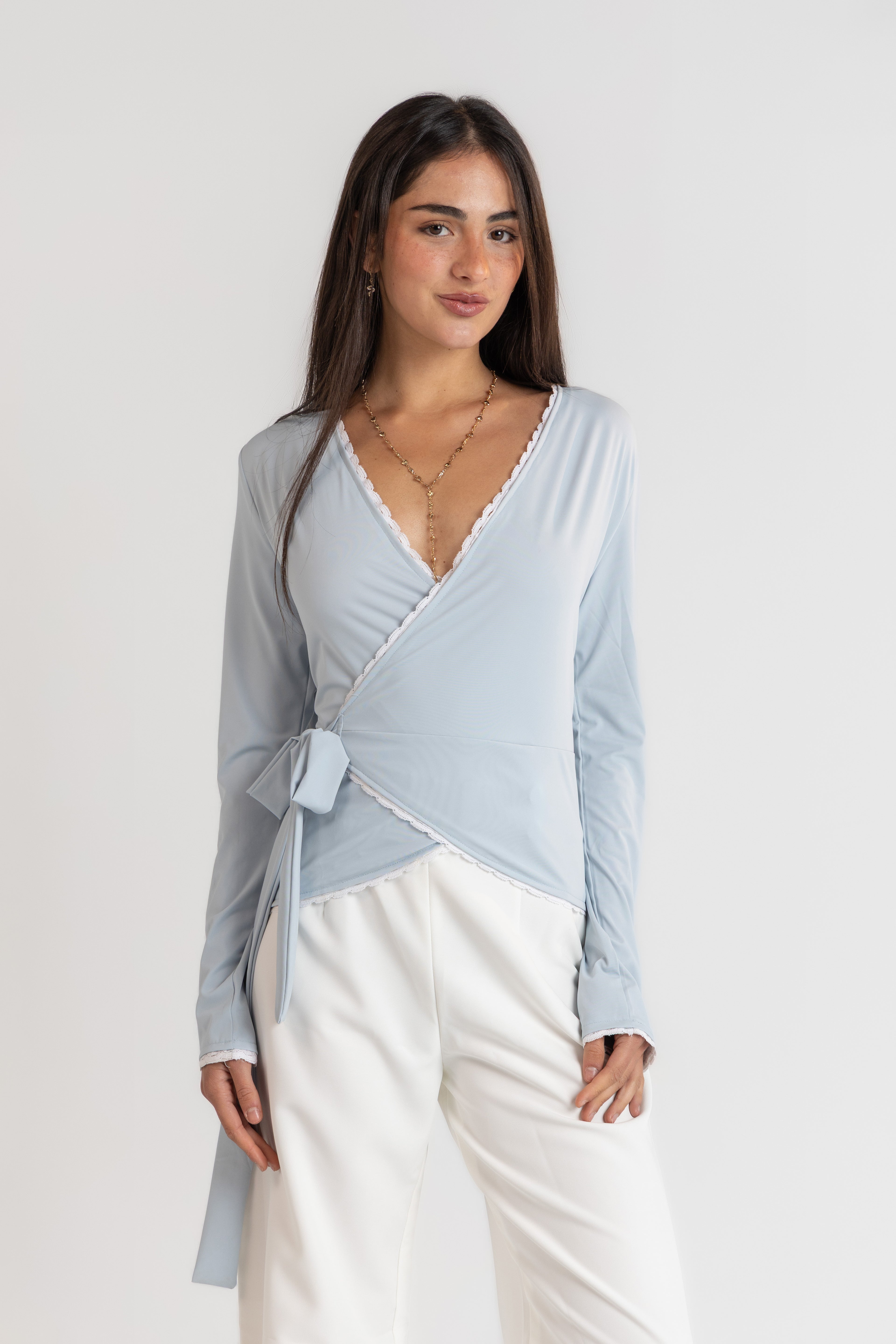 Woman wearing a light blue wrap top and white pants on a plain background