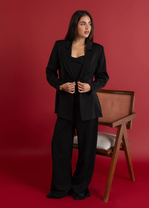 Woman in a black suit standing against a red background with a chair visible.